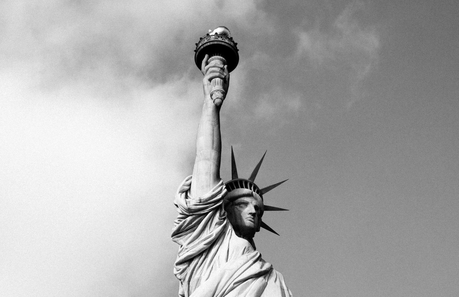The Statue of Liberty against a clouded sky, torch raised. Black and white.